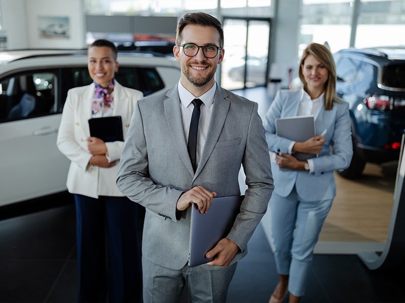 Three professional car dealership employees are smiling in a showroom